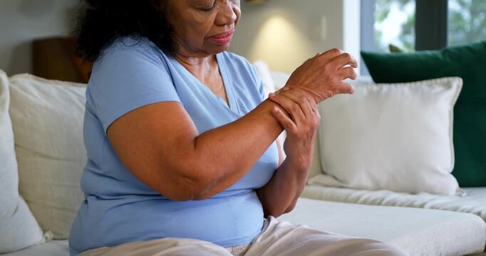 Senior African American woman raising right hand, rubbing right wrist for pain on sofa with pillows