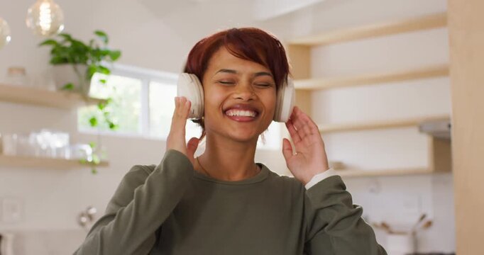 Adult woman placing white over-ear headphones while music starts, smiling and dancing in kitchen