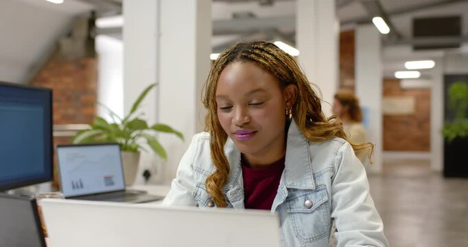 Female worker in denim jacket glancing at laptop leaning in studying at office as colleague passing