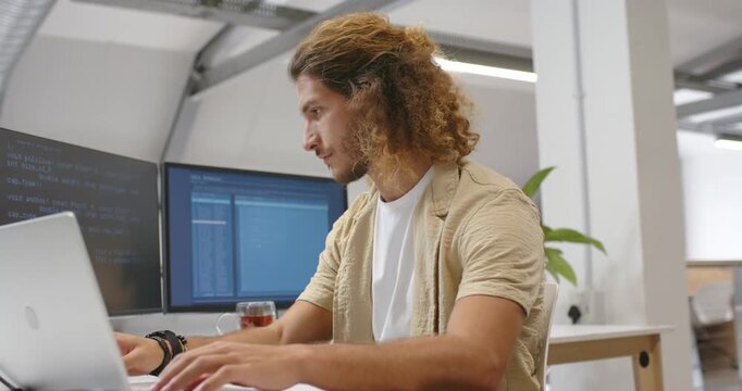 Adult male coder typing seeing code on left monitor pointing and tuning laptop debugging in office