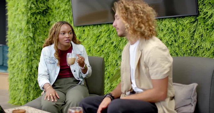 Diverse coworkers, woman raising latte mug and speaking, causing man turning and gesturing on sofa