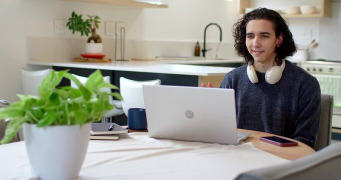African American man sitting at round kitchen table with headphones smiling, waving on laptop call