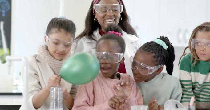 African American teacher in labcoat guiding kids placing green balloon on bottle showing gas at lab