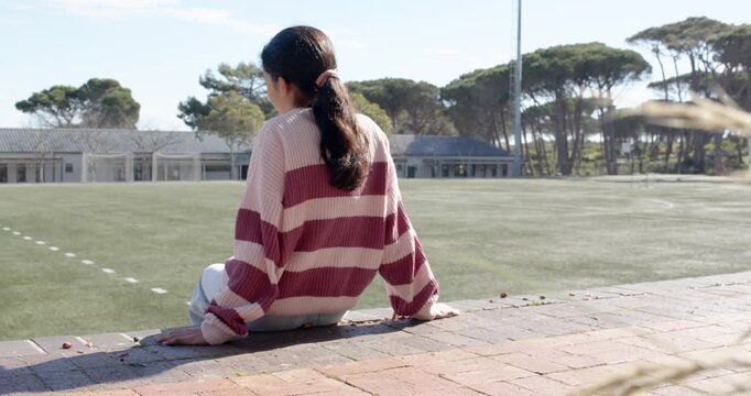 African American teenage female sitting on brick ledge on turf, noticing lens, turning and smiling
