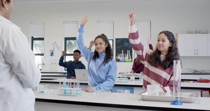 Female teens raising hands while teacher in lab coat asking question at bench, blue test tubes