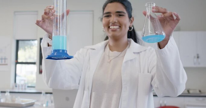 Labcoat woman holding cylinder and flask blue-liquid at bench, showing marks while camera nearing