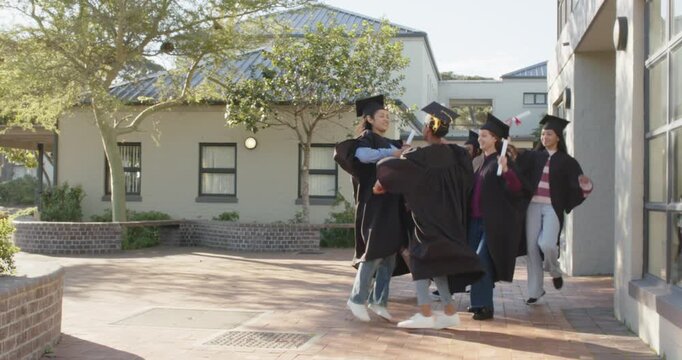 Diverse grads in gowns emerging from door waving diplomas and hugging in brick courtyard for grad