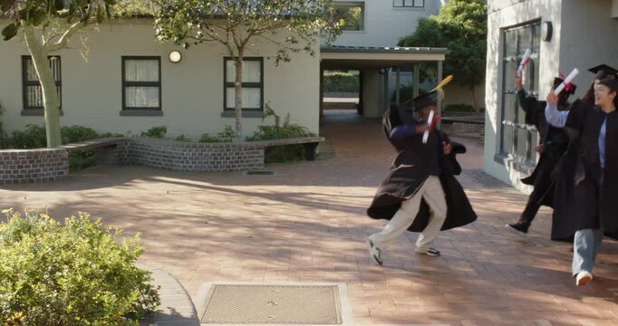 Diverse teenage peers celebrating grad entering right, dancing in gowns and waving diplomas on quad