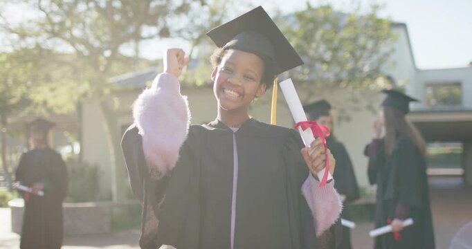 Youth African American female graduate smiling raising fist, hugging diploma after ceremony in cap