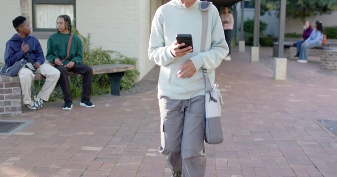 Teenage male entering, walking toward camera in brick plaza checking phone and adjusting sling bag