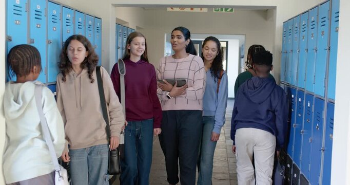 Diverse teen female classmates walking between classes at school past blue lockers holding tablet