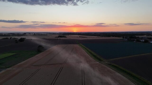 Golden light bathes the horizon above vast fields. Dust trails follow a tractor's path through cultivated earth. Sky fades from warm orange to soft lavender. Farmland stretches, bordered by green