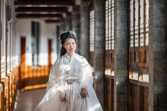 A female adult in white traditional hanfu stands in an ancient stone-and-wood corridor, with natural light casting soft shadows.
