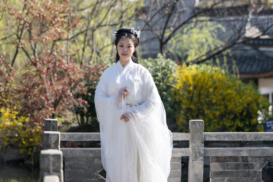 A young woman in white traditional hanfu stands on a stone bridge in a garden with green and red foliage, under daylight.