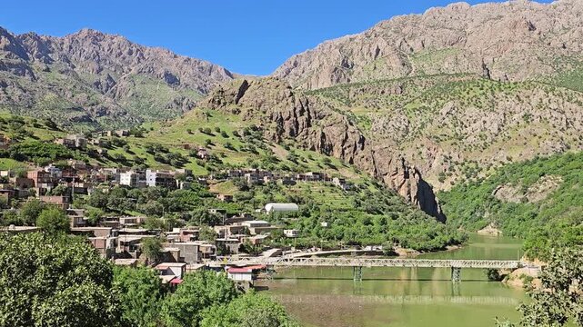 Sun-drenched mountain village nestled on a lush hillside overlooks a serene green river crossed by a bridge, framed by rocky peaks under a spring sky.
📍Selin village, Hawraman Takht, Kurdistan 