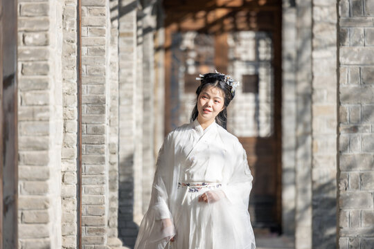 A young asian woman in white traditional hanfu stands in a sunlit ancient brick corridor, wearing decorative hairpins.