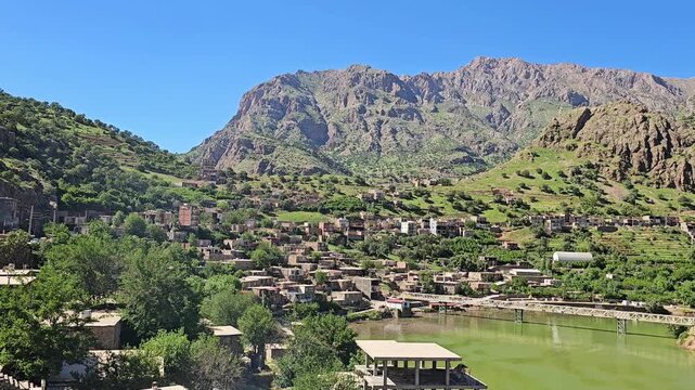 Sun-drenched mountain village nestled on a lush hillside overlooks a serene green river crossed by a bridge, framed by rocky peaks under a spring sky.
📍Selin village, Hawraman Takht, Kurdistan 