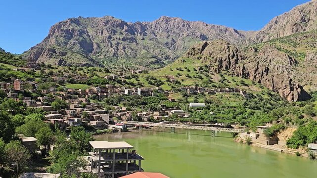 Sun-drenched mountain village nestled on a lush hillside overlooks a serene green river crossed by a bridge, framed by rocky peaks under a spring sky.
📍Selin village, Hawraman Takht, Kurdistan 
