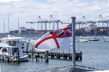 Traditional Royal Navy-style White Ensign flying aboard the historic HMAS Castlemaine, with moored yachts and industrial shipping cranes in the background, Williamstown, Melbourne, Australia © Doublelee