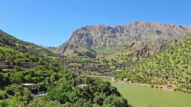 Tranquil green river winds beside a lush hillside village, framed by rocky spring peaks and orchard-covered slopes under a clear blue sky.
📍Selin village, Hawraman Takht, Kurdistan, Iran 