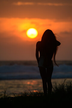 Silhouette of a young woman standing on a grassy shoreline, watching a brilliant sunset over the ocean as golden light reflects across the waves