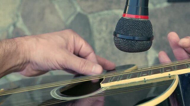 POV (slow zoom out): man playing acoustic guitar in front of a microphone. A guitar player in front of a microphone. Guitarist strumming a guitar. Artist plucking a guitar. Musician playing guitar