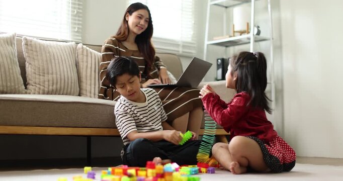 Asian mother working on a laptop from home, while her young son and daughter drawing at a coffee table while raising hands, signifying active child education and creative homeschooling