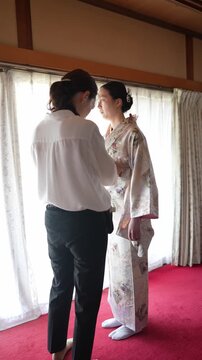 Soft light spills through window curtains. Woman gently adjusts kimono on standing bride. Helper wears simple white shirt and dark trousers. Red carpet contrasts with traditional wooden beams