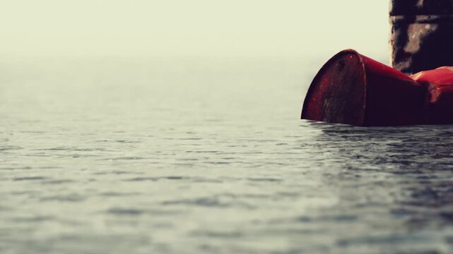 A red boat is half submerged in calm water. The scene shows a stone pier in the background. The setting is early morning with soft light reflecting on the water surface.