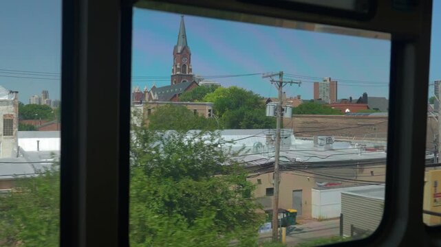 A church tower rises above trees and buildings in a Chicago neighborhood viewed from a moving train. The scene shows urban architecture, greenery, and everyday city life.