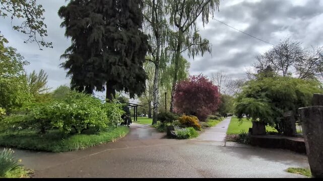 Video of a peaceful park entrance with a circular concrete gateway and three walking paths leading through green grass, colorful trees, and landscaped bushes on an overcast day.