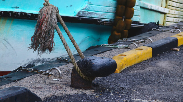 Frayed Mooring Rope Tied to Rusty Bollard on a Concrete Dock with a Teal Boat Hull in the Background