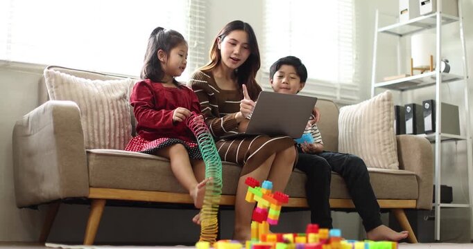 Asian mother working on a laptop from home, while her young son and daughter drawing at a coffee table while raising hands, signifying active child education and creative homeschooling