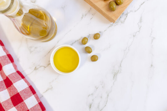 White marble surface holds olive oil and green olives. Red and white checkered cloth adds rustic charm. Glass bottle and bowl display golden oil, fresh. Olives rest near a wooden cutting board