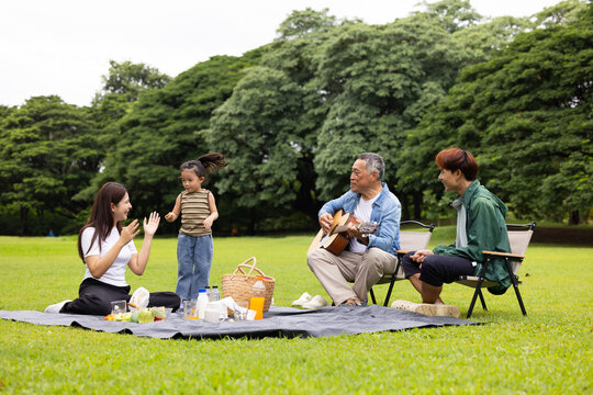 GrandFather playing acoustic guitar for child during picnic. Asian family enjoy outdoor picnic with food and drinks in park sunny day, outdoor activitie with daughter father, mother grandfather