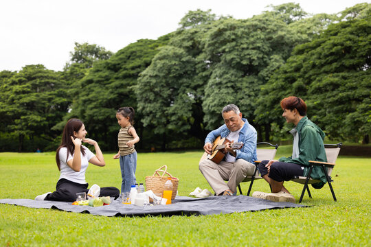 GrandFather playing acoustic guitar for child during picnic. Asian family enjoy outdoor picnic with food and drinks in park sunny day, outdoor activitie with daughter father, mother grandfather