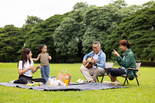 GrandFather playing acoustic guitar for child during picnic. Asian family enjoy outdoor picnic with food and drinks in park sunny day, outdoor activitie with daughter father, mother grandfather