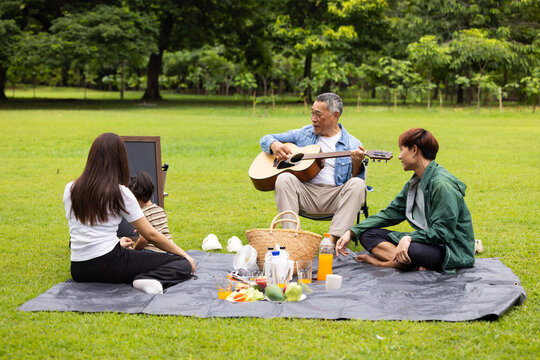 GrandFather playing acoustic guitar for child during picnic. Asian family enjoy outdoor picnic with food and drinks in park sunny day, outdoor activitie with daughter father, mother grandfather