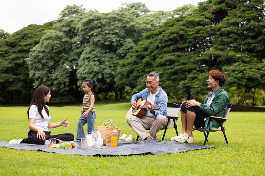 GrandFather playing acoustic guitar for child during picnic. Asian family enjoy outdoor picnic with food and drinks in park sunny day, outdoor activitie with daughter father, mother grandfather