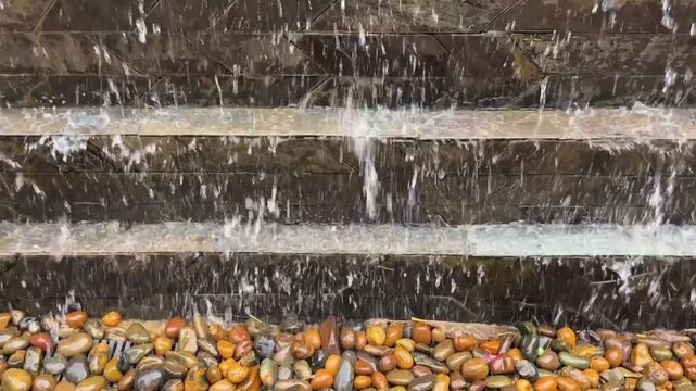 Streams of water flow down a multi-level stone wall, forming a decorative cascade. Below, wet river pebbles form the drainage area. A backdrop for design, architecture, and landscape.