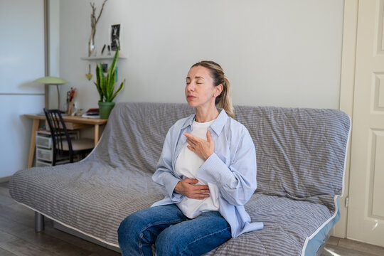 Calm mature woman do stress relief deep breathing self helping therapy, female sitting on sofa holding hands on chest inhaling air, focusing on physical and mental relaxation, healing zen practice