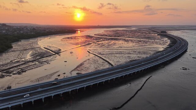 Aerial sunset view of the Nusa Dua toll bridge curving over coastal mudflats and shallow water, with golden sky reflections and sparse traffic highlighting coastal infrastructure and travel atmosphere