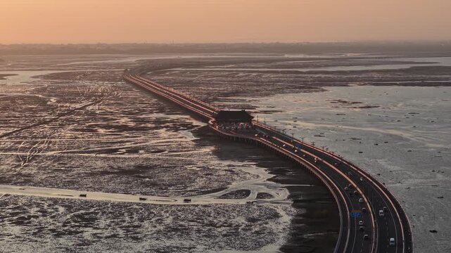 Aerial sunset view of the Nusa Dua toll bridge and coastal causeway stretching across tidal mudflats, with evening traffic lines reflecting golden light, highlighting infrastructure, travel