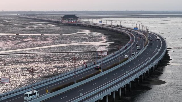 Aerial view of a long curved causeway bridge and toll gate carrying cars and motorcycles over expansive tidal flats at dusk, conveying travel, infrastructure, coastal landscape and quiet atmosphere
