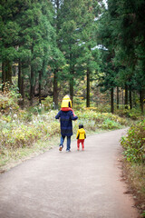 Fototapeta premium Rear view of a father carrying one child on his shoulders and walking down a quiet forest path with his children in late autumn.
