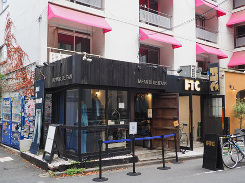 TOKYO, JAPAN - July 31, 2026: Japan Blue Jeans store in Shibuya. Next to it are some drinks vending machines and FTC skateboard store. The stores are on the first floor of a 1960s apartment building.
