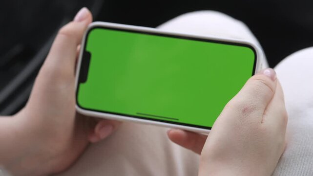 Close-up of hands holding smartphone with green screen. Fingers tap screen, showing interaction. Person appears focused, relaxed, engaged with device. Ideal for app demos, social media