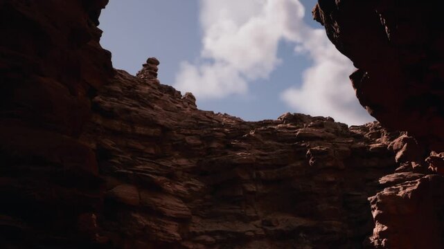 Two large rock walls frame a narrow opening. Blue sky shines above, with clouds floating. Sunlight highlights the rugged texture of the rocks.