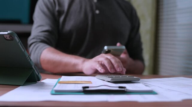 Man holding smartphone calculates personal budget inside home office, analyzing financial documents, credit card details, tablet screen data, managing household expenses, planning saving goals.