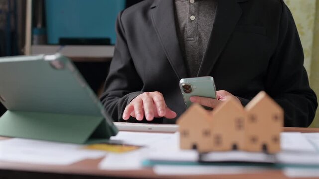 Male agent holding mobile phone types computer keyboard near tablet, analyzing real estate investment options near small wooden house models inside workspace, planning financial budget.